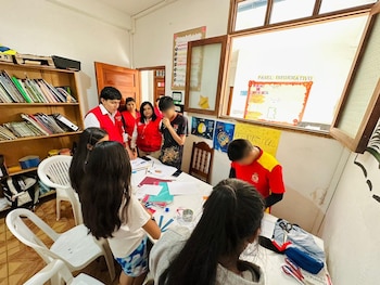 Varias personas, incluidos adultos con chalecos rojos y niños, se agrupan alrededor de una mesa blanca con papeles en una habitación con estanterías y paneles informativos