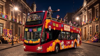 Autobús rojo de dos pisos decorado con flores de cempasúchil y calaveras, guiado por un hombre con micrófono junto a pasajeros maquillados de noche.