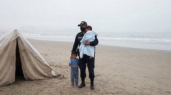 Policía peruano sostiene niño en manta azul junto a otro niño pequeño en playa brumosa. Carpa y mar visible al fondo con cielo gris claro.