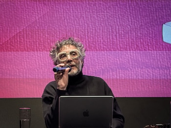 Hombre con pelo rizado gris, barba y gafas claras, habla por micrófono. Delante, un portátil y un vaso de agua. Al fondo, una pantalla digital con tonos rosas