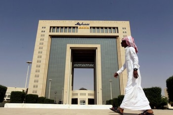 Hombre con vestimenta tradicional saudí camina frente a la imponente sede de SABIC, un edificio moderno beige con grandes ventanales de vidrio, bajo un cielo azul despejado