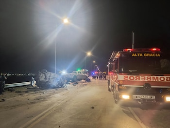 Un camión de bomberos estacionado de noche en una vía al lado de un automóvil volcado y destruido, con personal trabajando de fondo