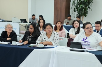 Primer plano de un grupo de mujeres sentadas en mesas con tazas, cuadernos y bolígrafos. Otras personas están sentadas en el fondo
