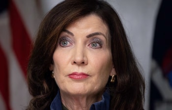 Primer plano de una mujer con cabello castaño oscuro, ojos azul verdosos, labios rojos y pendientes dorados, contra un fondo borroso de la bandera de EE. UU