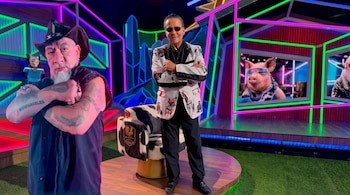 Dos hombres de pie en un estudio de televisión con luces de neón, un sillón con estampado de vaca y pantallas al fondo.