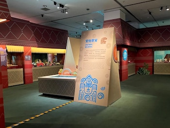 Interior de una exposición cultural con paredes rojas y patrones geométricos, vitrinas con artefactos, un panel informativo central y suelo verde con línea de advertencia