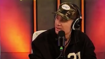 Hombre usando gorra de camuflaje, auriculares negros y chaqueta oscura con el número veintiuno, habla en un micrófono en un fondo con luces rojas y naranjas