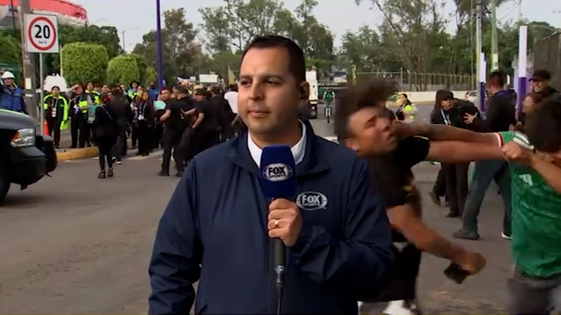Mientras un reportero de Fox Sports transmite en vivo, dos aficionados se agreden físicamente en el fondo, con una multitud de personas y un vehículo visible en la calle.