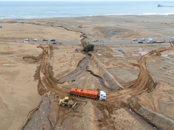 Vista aérea de carretera cortada por erosión, camión con carga naranja, retroexcavadora amarilla, vehículos y personas. Al fondo, el mar y la costa desértica