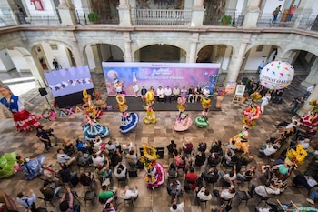 Vista aérea de un patio colonial con un escenario donde autoridades presentan La Guelaguetza 2026, rodeadas de bailarines con trajes típicos y un público observando