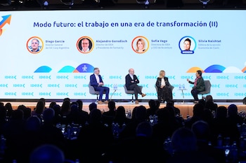 Cuatro personas sentadas en un escenario frente a una pantalla con sus nombres y rostros ante un público en la oscuridad