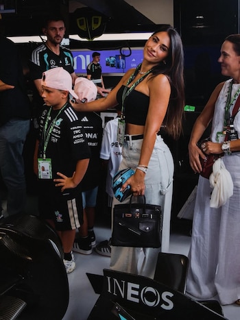 Antonela Roccuzzo con top negro y jeans claros sonríe en un garaje de F1, sujetando un bolso negro y un objeto azul. Niños y adultos la rodean, algunos con ropa de Mercedes F1