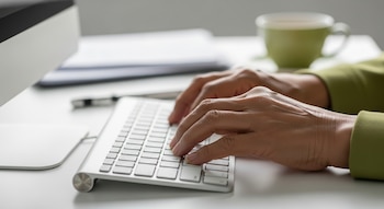 Primer plano de manos adultas mayores de rasgos asiáticos tecleando en un teclado de computadora moderno. Un escritorio blanco ordenado, con papeles y una taza de té desenfocados detrás.