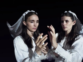 Dos mujeres con cabello oscuro y coronas de flores blancas con velos, vestidas con trajes blancos, se miran con las manos levantadas frente a un fondo negro