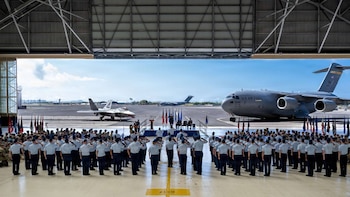 Un grupo de militares en formación en un hangar, con un avión de transporte U.S. Air Force y un caza en la pista. Al fondo, más aeronaves y una ciudad