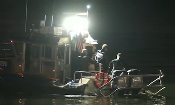 Barco oscuro de la policía con insignia NYPD iluminado por una luz potente en el agua. Tres personas con uniformes oscuros a bordo, bandera de Estados Unidos y salvavidas