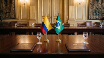 Dos banderas de escritorio de Colombia y Brasil sobre una mesa de madera con carpetas de cuero y copas con agua en un salón clásico.