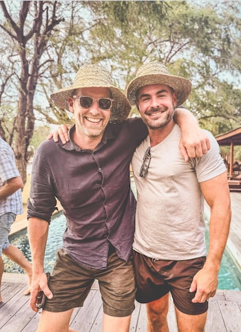 Dos hombres sonrientes, incluido Zac Efron, con sombreros de paja y ropa casual, posan al aire libre con una piscina y árboles de fondo