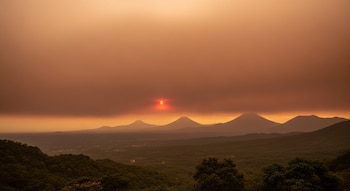 El sol rojizo se asoma en un cielo anaranjado, sobre siluetas de montañas y una extensa zona de vegetación y árboles en primer plano.