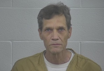 Retrato frontal de un hombre de mediana edad con cabello castaño y ojos claros, vestido con una camisa marrón y una camiseta blanca, frente a una pared de ladrillo gris