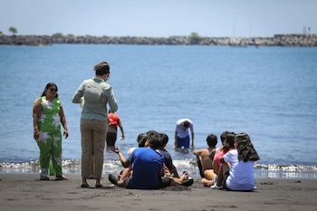 Varias personas sentadas y de pie sobre una playa de arena negra observan el mar con un rompeolas al fondo