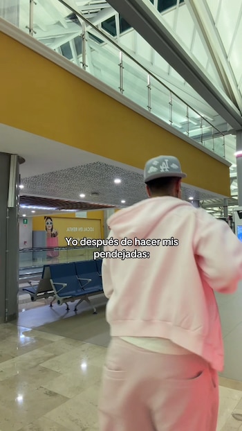 Un hombre de espaldas viste una sudadera de color rosa y una gorra gris mientras camina por el pasillo de un aeropuerto con bancas azules