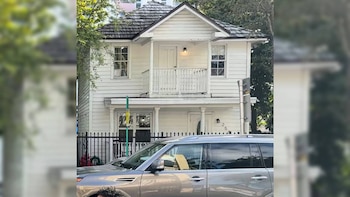 Una casa blanca de dos pisos con un balcón superior. Un SUV plateado Land Rover está estacionado parcialmente frente a ella, detrás de una valla negra