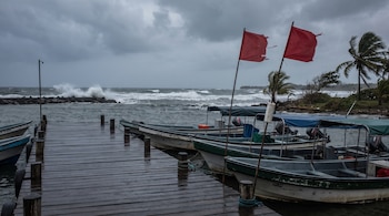 Muelle de madera mojado con varias embarcaciones pequeñas amarradas, dos banderas rojas rasgadas, mar agitado con olas grandes y cielo muy nublado.
