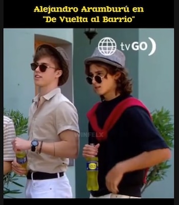 Dos jóvenes con sombreros y gafas de sol posan en el exterior; uno, con cabello largo, viste una camiseta oscura y un suéter rojo, el otro lleva camisa beige. Ambos sujetan botellas de bebida amarilla
