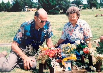 Hombre y mujer mayores arrodillados en un cementerio junto a una tumba con flores y una foto enmarcada. El hombre usa gafas de sol
