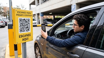 Un hombre dentro de un automóvil gris apunta su teléfono inteligente hacia un cartel amarillo con un código QR en un estacionamiento.