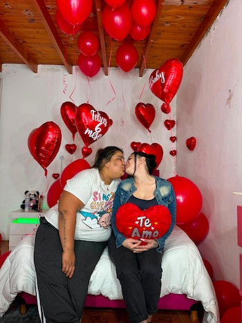 Dos mujeres se besan en una cama. El fondo muestra una habitación decorada con globos rojos, algunos con forma de corazón y texto. Una mujer sostiene un cojín rojo
