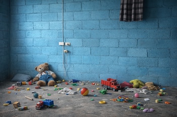 Imagen de un cuarto desordenado con paredes de ladrillo azul. Un oso de peluche y numerosos juguetes, incluyendo camiones y bloques, están esparcidos en el suelo.
