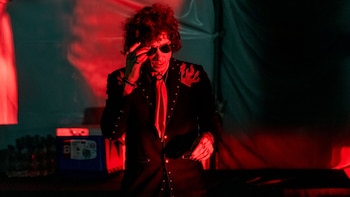Bunbury, con cabello rizado oscuro y gafas de sol, ajusta sus gafas mientras viste una chaqueta negra con llamas rojas, bajo una luz roja intensa en el escenario
