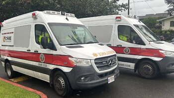 Dos ambulancias blancas con franjas rojas y luces de emergencia; la frontal muestra el logo del departamento de servicios de emergencia de Hokulū y texto invertido