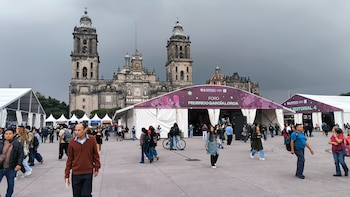 Varias personas caminan frente a carpas blancas con carteles de eventos culturales en una gran plaza, con una catedral antigua al fondo