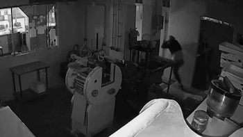 Foto en blanco y negro de un interior industrial con maquinaria. Una persona de espaldas está en movimiento cerca de una puerta y cajas apiladas