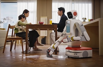 Un robot blanco y rojo limpia un derrame en un suelo de madera. Al fondo, una familia de tres personas sentada en una mesa de madera con sillas y un bol de frutas