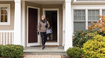 Mujer con gorro, chaqueta acolchada, guantes y mallas negras, con una bolsa de deporte, sale por la puerta principal de una casa. Hay arbustos y ventanas.