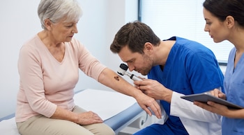 Un doctor examina una lesión cutánea en el antebrazo de una mujer mayor con un dermatoscopio, mientras una asistente anota en una tableta.