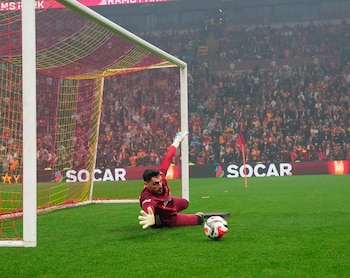 Un portero con uniforme rojo se lanza en el césped verde para atajar un balón de fútbol blanco y rojo, con una portería y una multitud en el fondo