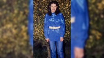 Una mujer con cabello oscuro y suéter azul estampado con motivos geométricos y cinturón blanco posa de cuerpo entero frente a un follaje verde oscuro