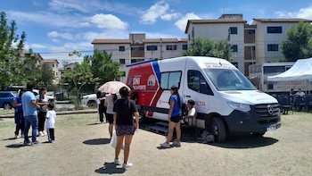 Una unidad móvil de identificación con rótulos rojos y azules. Personas de pie cerca de la furgoneta y una carpa blanca con sillas. Edificios y árboles al fondo