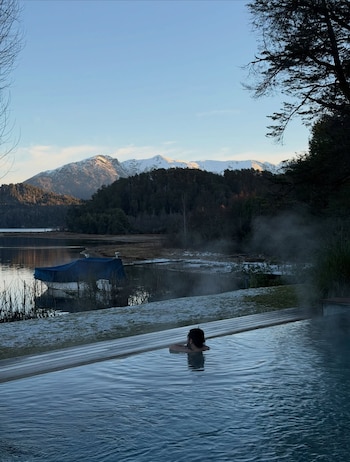 Persona sumergida en piscina con bordes de madera, vapor sobre el agua, lago, bote cubierto, árboles y montañas nevadas en el horizonte