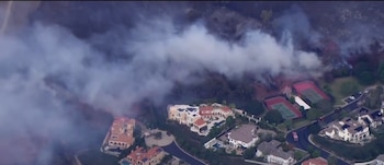 Vista aérea de una densa columna de humo sobre una colina junto a casas residenciales, calles y canchas de tenis
