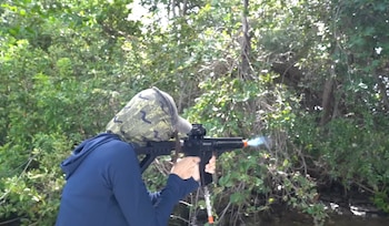 Una persona cubierta con una capucha de camuflaje dispara una réplica de arma de aire comprimido frente a follaje denso