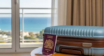 Una maleta azul claro y un pasaporte británico color borgoña sobre una mesa, con una ventana que muestra el mar y cortinas claras al fondo.