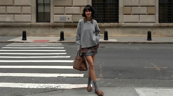 Una mujer sonríe y posa de pie en la calle, con un suéter gris, falda y un bolso de cuero marrón. Se ve un cruce peatonal y un edificio de piedra detrás.