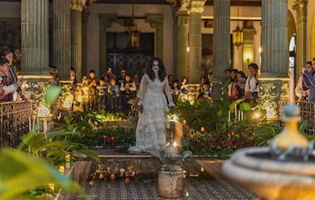 Persona disfrazada de figura fantasmal en vestido blanco con maquillaje pálido en un patio colonial iluminado. Multitud observa desde balcones entre columnas y vegetación