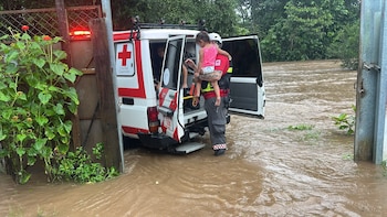 Tres rescatistas con uniformes de la Cruz Roja, cascos y chalecos, junto a un vehículo de rescate blanco. El fondo muestra un paisaje montañoso y húmedo con vegetación